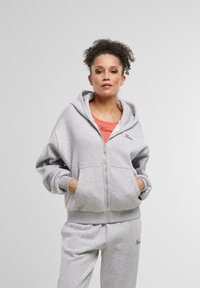 Sivý zipsový sveter s prednými vreckami a rebrovanými manžetami, v kombinácii so sivými teplákmi s malým vyšívaným logom.