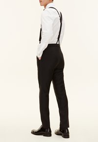 Schwarze Hose mit Hosenträgern und einem weißen Hemd mit Knöpfen. Glatter, maßgeschneiderter Stoff; schimmernde schwarze Lackschuhe runden das Outfit ab.