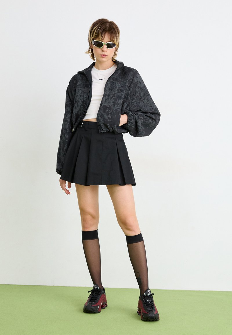 Veste bomber à motifs floraux noirs, top court blanc, jupe plissée noire, chaussettes montantes noires à rayures, baskets bordeaux et noires.