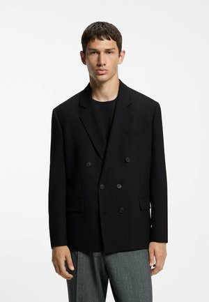 Junger Mann trägt schwarzen zweireihigen Blazer mit schwarzen Knöpfen und grauen gemusterten Hosen, steht vor einem schlichten weißen Hintergrund.