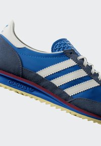 Sinine ja tumesinine Adidas toss valgete triipude, valgete paelte, tekstuurse sinise keele ja kollase tallaga, millel on Adidas logo.