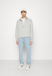 Sudadera de forro polar gris con media cremallera y logo azul. Combinada con vaqueros azul claro y zapatillas blancas. Gorro beige neutro.