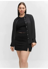 Čierny priliehavý crop top spárovaný s čiernou mini sukňou s bočným rozparkom. Veľké košeľové tričko, tiež čierne, nosené otvorené cez outfit.