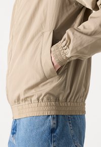 Beige letvægtsjakke med elastiske manchetter og talje, med en sidelomme og glat tekstur. Sammen med blå denim jeans.