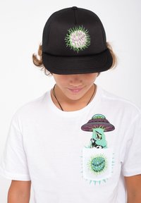 Musta trucker-hattu vihreällä graafisella logolla, yhdistetty valkoiseen t-paitaan, jossa on alien-graafika taskussa ja kirkkaat yksityiskohdat.