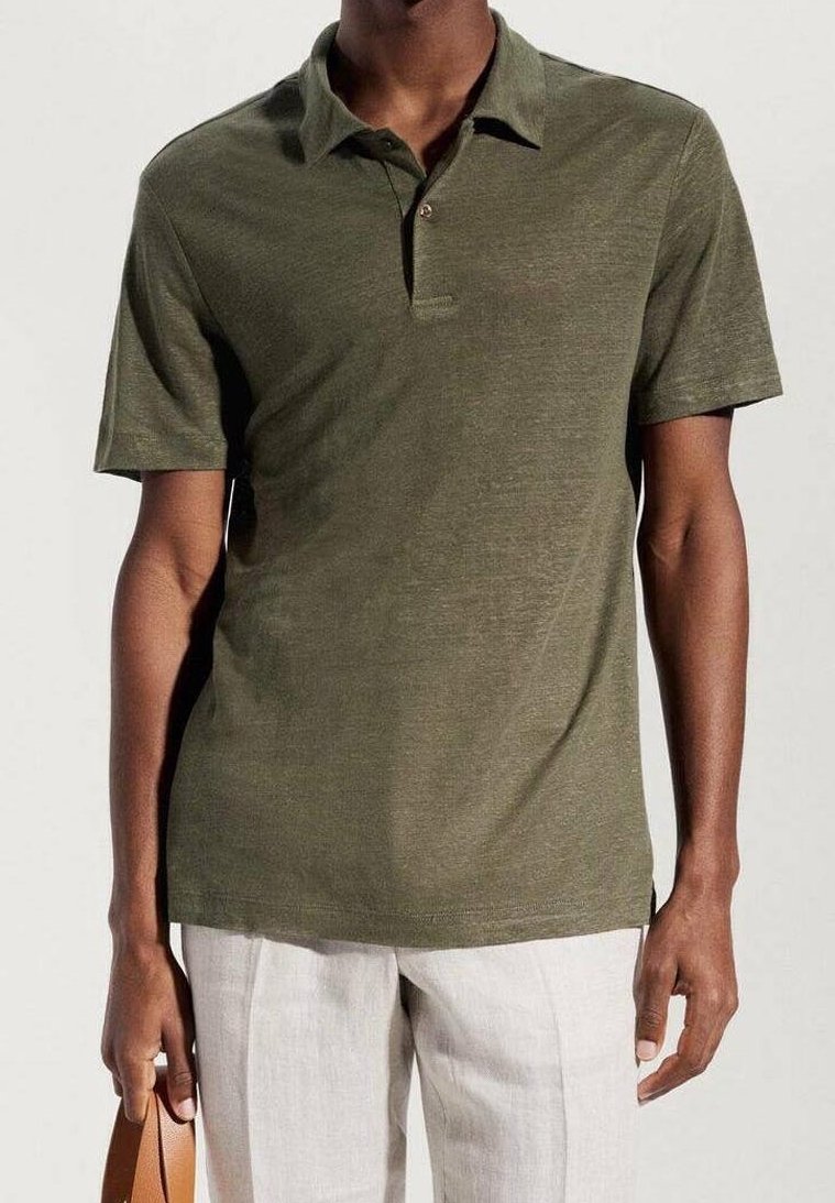 Homme portant un polo à manches courtes vert olive et un pantalon beige clair, tenant une poignée de sac en cuir marron dans la main droite.