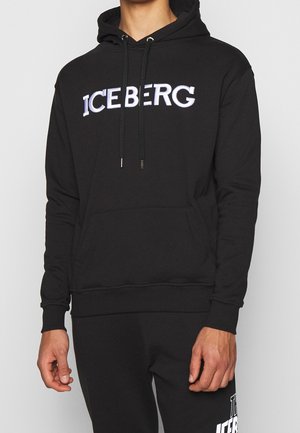 Mężczyzna w czarnej bluzie z kapturem z białym napisem "ICEBERG" na klatce piersiowej oraz czarnych spodniach z widocznym częściowo białym napisem.