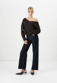 Femeie îmbrăcată cu un pulover închiset, cu umerii la vedere, de culoare maro închis, blugi largi cu talie înaltă, de culoare închisă, tocuri slingback negre și purtând o geantă de umăr neagră.