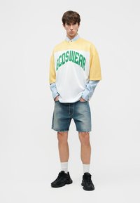 Νέος άνδρας φορώντας κίτρινο και λευκό πουκάμισο GCDSWEAR πάνω από μπλε ριγωτή μακρυμάνικη μπλούζα, τζιν σορτς, λευκές κάλτσες και μαύρα αθλητικά παπούτσια, στέκεται με τα χέρια στις τσέπες.