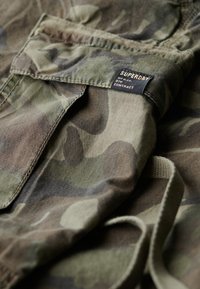 Camo-gepatronneerde stof in tinten groen en bruin, met een zwart label met de tekst "SUPERDRY" en een opgevouwen pocketdetail.