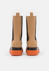 Botas de tornozelo castanhas e pretas com um acabamento texturizado, apresentando uma sola chunky laranja e painéis laterais elásticos para facilitar o calçar.