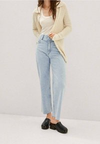 Cardigan en maille beige porté sur un débardeur blanc, associé à un jean taille haute bleu clair et des sabots noirs ornés de clous décoratifs.