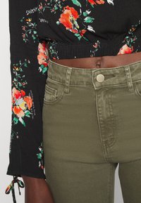 Top preto floral com mangas longas, apresentando padrões em vermelho e verde, combinado com calças verdes-oliva de cintura alta com fecho de botão frontal.