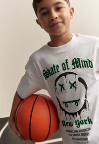 Witte katoenen T-shirt met een groen en zwart ontwerp met de tekst "Skate of Mind" en een schedelmotief. Persoon houdt een oranje basketbal vast.