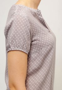 Nærbillede af en person iført en lyse lilla kortærmet skjorte med små hvide blomsterprint og elastiske kanter på ærmerne.