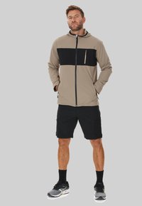 Leichte beige und schwarze Zip-Jacke mit Kapuze, ausgestattet mit einer Brusttasche; kombiniert mit schwarzen Shorts und Sportschuhen.