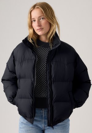 Marineblaue Steppjacke mit hohem Kragen, gestepptem Design, Reißverschluss vorne und elastischen Bündchen. Getragen über einem gemusterten Pullover und blauen Jeans.