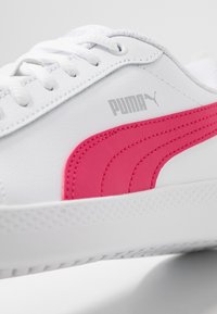 Biała sneaker Puma z jasnoróżowym paskiem po boku i białymi sznurowadłami, ukazująca teksturowaną podeszwę i logo marki blisko górnej krawędzi.