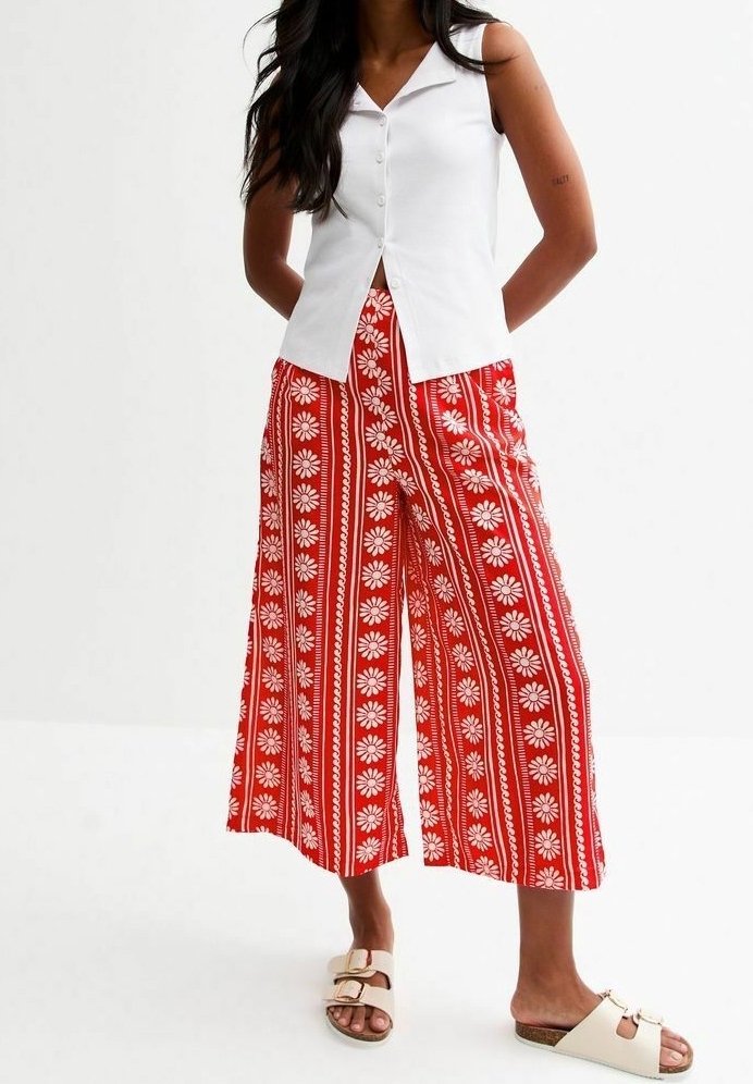 Femme portant un chemisier blanc sans manches à boutons, un pantalon large rouge à rayures florales blanches, et des sandales beiges à boucle.