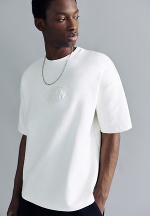 Ung mand iført en hvid oversized t-shirt med korte ærmer og et sølvkædehalskæde, kigger til venstre mod en ensfarvet baggrund.