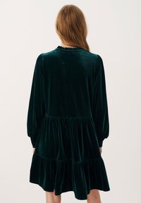 Abito verde in velluto con scollatura arricciata e maniche lunghe a sbuffo. Presenta una gonna a strati e una texture liscia e morbida.