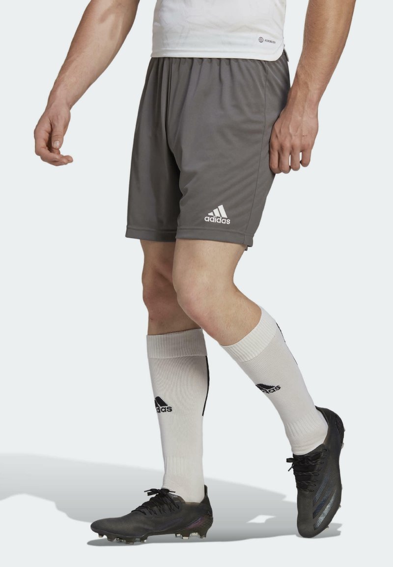 Homme portant un short gris Adidas, des chaussettes blanches Adidas montantes et des crampons de football noirs, debout devant un fond uni.