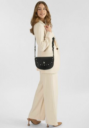 Zwarte leren crossbodytas met zilveren studs, afgeronde vorm, verstelbare riem, met een logodetail aan de voorkant, gedragen bij een licht beige outfit.