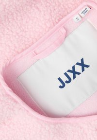 Blødt lyserødt struktureret stof med syet hvidt mærke med teksten "JJXX" og et mindre mærkat, der angiver størrelse small, lavet i Kina.