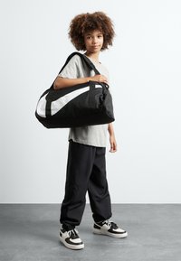 Black duffle bag with white curved stripe, sturdy fabric, large shape, adjustable shoulder strap, and double handles.