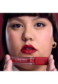 Rouge à lèvres liquide mat dans un tube transparent avec un bouchon rouge, tenu dans une main ; le modèle présente une couleur de lèvres rouge vif et un maquillage neutre contre un fond uni.