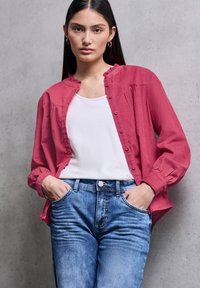 Červená košile na knoflíky s volánkovým límcem přes bílý tílko, doplněná modrými džíny s vysokým pasem, které mají přední kapsy.