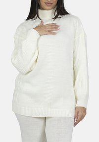 Pull en tricot couleur crème avec col roulé, motifs texturés, épaules tombantes et poignets côtelés. Porté avec un pantalon assorti texturé.