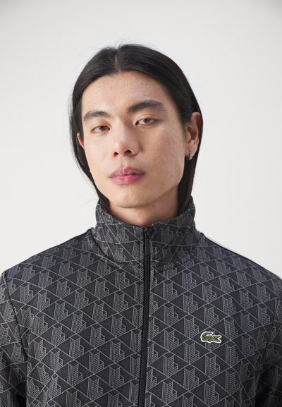 Veste zippée noire avec un motif géométrique, un col haut et un accent de logo vert. Fabriquée en tissu texturé avec une coupe près du corps.