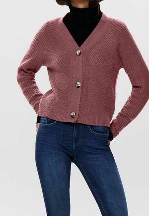 Femme portant un cardigan en maille rose poudré boutonné sur un col roulé noir avec un jean bleu, les mains dans les poches arrière, debout devant un fond blanc.