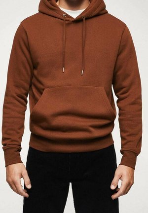 Persona con sudadera marrón con capucha, bolsillo frontal y cordones, combinada con pantalones negros, sobre un fondo liso.