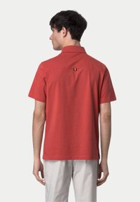 Chemise à manches courtes avec col, en coton rouge uni, avec un petit détail jaune et noir au dos. Texture lisse et coupe décontractée.