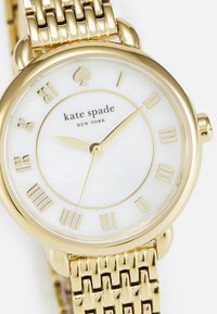 Guldtonad klocka med rund boett, vit pärlemorsfärgad urtavla, romerska siffror och ett vävt metallarmband. Har en logotyp i mitten.