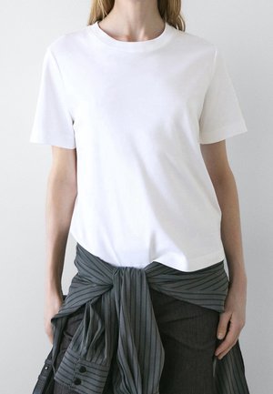 Person wearing a plain white short-sleeve t-shirt with a dark pinstriped shirt tied around their waist against a plain background.