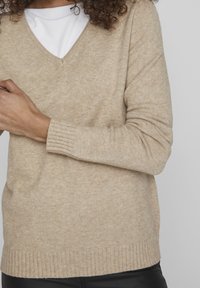 Beige tröja med v-ringad design, ribbad manschetter och strukturerad stickning. Bärs över en vit skjorta, med mjukt tyg och avslappnad passform.