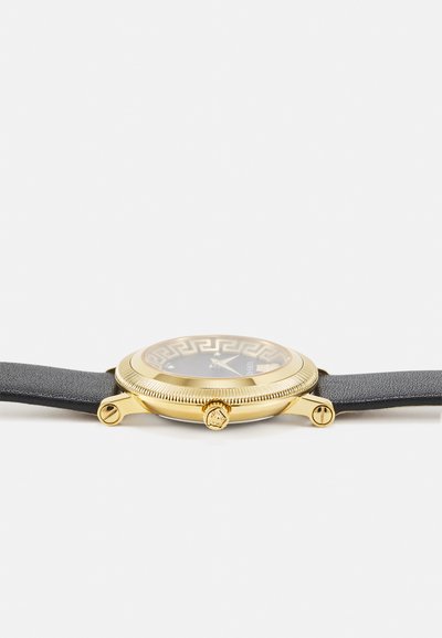Gold-tone wristwatch with a round case, black textured leather strap, patterned black dial, and crown featuring a decorative accent.