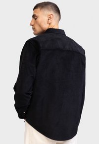 Camicia di velluto corduroy scura con colletto a bottone e maniche lunghe. Tessuto testurizzato ben visibile e orlo curvo.