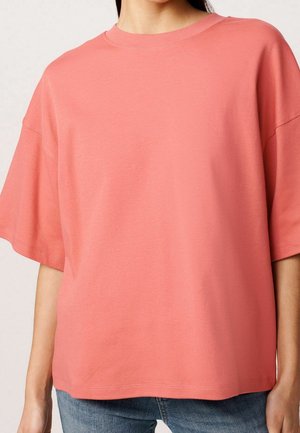 Vrouw met een loszittend, koraalroze T-shirt met korte mouwen, gecombineerd met een blauwe spijkerbroek, getoond van schouders tot heupen.