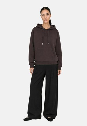 Donna con capelli scuri raccolti, indossa una felpa con cappuccio marrone scuro, pantaloni neri a gamba larga e scarpe nere, in piedi davanti a uno sfondo chiaro e uniforme.