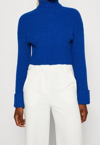 Korte blauwe ribgebreide coltrui met lange mouwen en brede manchetten, gecombineerd met witte broek met hoge taille. Geaccentueerde textuur van de stof.