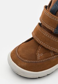 Braune Wildlederschuhe mit zwei Klettverschlüssen, runder Spitze, weißer Gummisohle und kontrastierenden Nähten. Nahaufnahme.