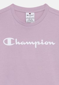 Camiseta de algodón de color púrpura claro con escote redondo. Presenta un destacado logo en cursiva en blanco de "Champion" en la parte frontal.
