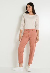 Haut beige à manches longues avec des boutons dorés, associé à un jean taille haute de couleur saumon et des baskets blanches. Design ajusté.
