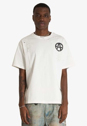 Giovane uomo con capelli corti che indossa una T-shirt bianca usurata con grafica nera sul petto e jeans chiari, in piedi davanti a uno sfondo bianco.