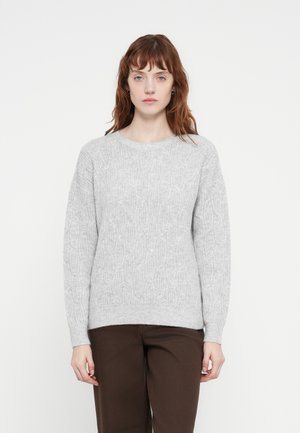 Hellgrau gestrickter Pullover mit einem strukturierten Zopfmuster, Rundhalsausschnitt und langen Ärmeln, kombiniert mit dunkelbraunen Hosen.