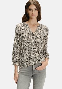 Kobieta w beżowej i czarnej wzorzystej bluzce V-neck z rękawami do trzech kwart, w jasnoszarych dżinsach, stojąca z jedną ręką w kieszeni.